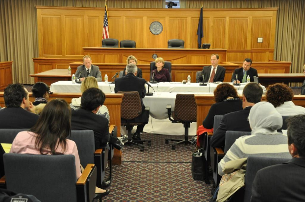 Ein Versammlungsraum mit Menschen auf Stühlen, zwei ausländische Flaggen, ein oranger Schreibtisch mit Wasserflaschen und ein Mikrofon.