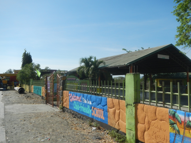 Schulgebäude mit einer Wandmalerei, umgeben von einem Zaun, einer Hütte, Bäumen, Fahrzeugen, Pfählen und einem bewölkten Himmel.