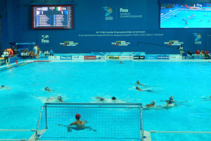 Eine Gruppe von Menschen spielt Wasserball in einem großen Pool mit einem Netz in der Mitte, Zuschauern auf der linken Seite und einer Wand mit Text und Logos im Hintergrund bei den FINA-Weltmeisterschaften 2016 in Kazan.
