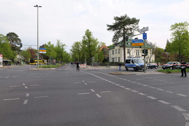 Ein Kreisverkehr mit einem Polizeiwagen am Straßenrand, fahrende Fahrzeuge, Fußgänger auf dem Gehweg, Verkehrsampeln, Bäume, Gebäude mit Fenstern und bewölkter Himmel.