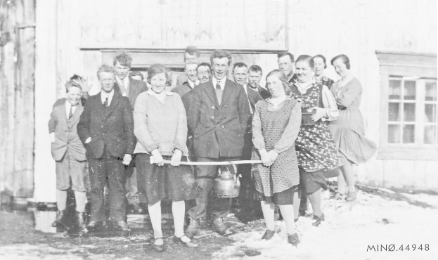 Schwarzes und weißes Foto von Menschen, die lächeln und ein Band halten, vor einem Schulgebäude mit einem Fenster und einer Tür.