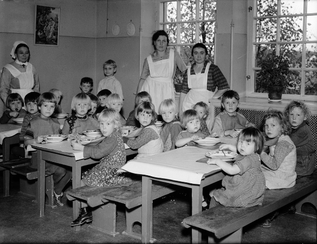 Kinder sitzen an Tischen in einem Klassenzimmer und essen von Tellern, mit Bänken rund um die Tische und einem gerahmten Foto an der Wand dahinter; Fenster rechts zeigen Pflanzen draußen.