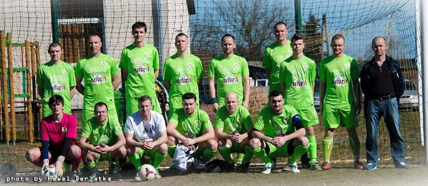 Eine Gruppe von Männern in grünen T-Shirts steht auf einem Fußballfeld, einige in einer Hockposition, mit einem Ball vor ihnen und einem Torpfosten rechts daneben, mit Bäumen, Gebäuden, Pfählen, Fahrzeugen und dem Himmel im Hintergrund.