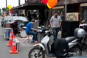 Eine Person, die Musikinstrumente im Freien spielt, mit Häusern, Bäumen, Menschen, Fahrzeugen und Luftballons im Hintergrund.