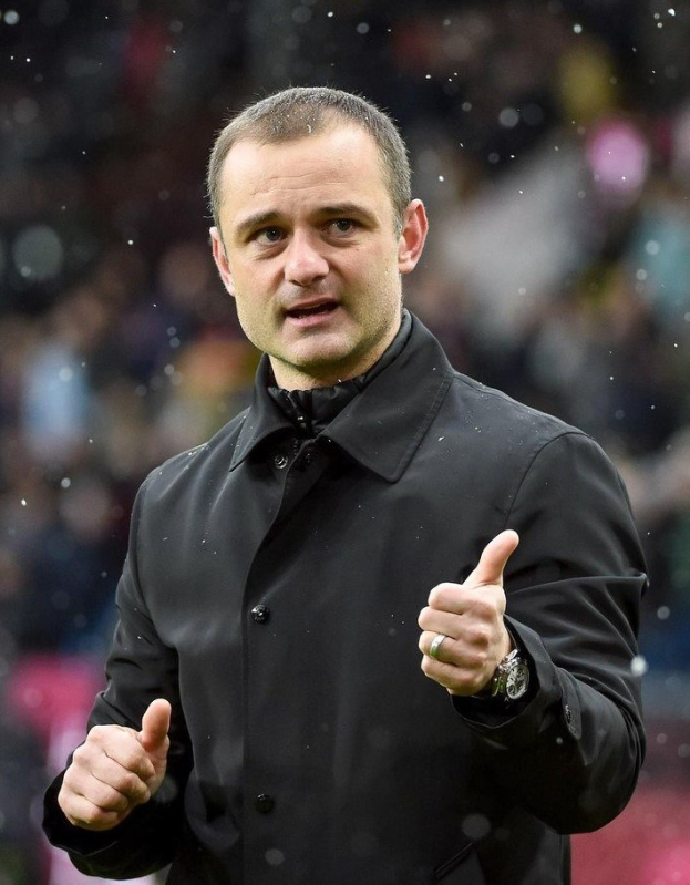 Mann in schwarzer Jacke zeigt Daumen hoch, trägt Uhr, mit unscharfem Hintergrund.