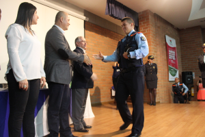 Eine Gruppe von Menschen auf einer B√ľhne mit einer Person in Uniform, die eine M√ľtze h√∂lt, einer Frau in einem wei√üem Hemd, einem Banner, einem Lautsprecher auf einem St√§nder, Vorh√§ngen, einem Tisch und einem Bildschirm an der Wand.