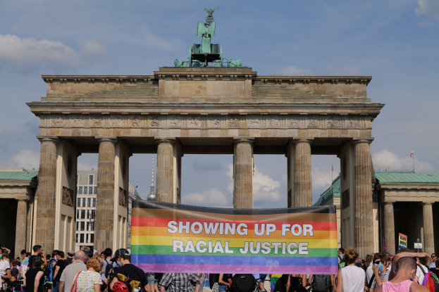 Eine Gruppe von Menschen mit einer "Racial Justice"-Schlagzeile vor dem Brandenburger Tor in Berlin, mit den Säulen und der Statue des Tors im Hintergrund und Gebäuden und bewölktem Himmel.