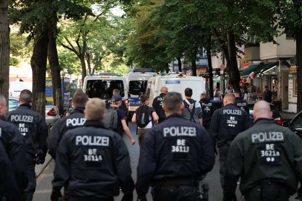 Polizisten gehen eine von Bäumen gesäumte Straße mit parkenden Fahrzeugen und Passanten entlang.