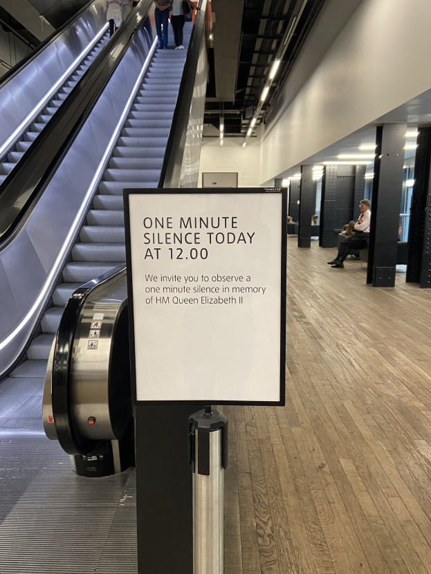 Eine Rolltreppe im Flughafen mit einem Schild, auf dem "Eine Minute Schweigen heute" steht, einige Menschen darauf und Deckenleuchten im Hintergrund.