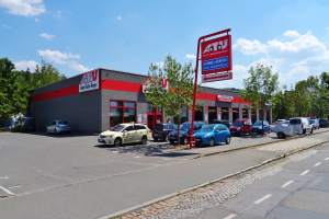 Autohaus mit parkenden Autos, umgeben von Gebäuden, Bäumen, Strommasten, Schildern und einer Straße unter einem bewölkten Himmel.