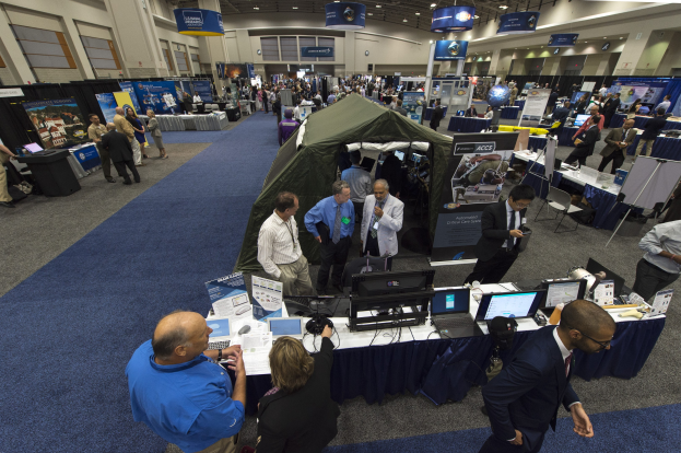 Große Gruppe von Menschen auf einer Karriere-Messe um Tische mit Laptops, Monitoren und Papieren versammelt, mit einem Zelt, Schildern, Deckenleuchten und Fenstern im Hintergrund.