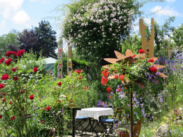 Ein Garten auf dem Chelsea Flower Show mit bunten Blumen und Pflanzen, einem Tisch und Stühlen in der Mitte, die mit einem Tuch bedeckt sind, umgeben von Bäumen unter einem bewölkten Himmel.