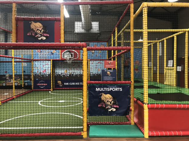 Große Indoor-Trampolinhalle mit einem Basketballfeld in der Mitte, umgeben von einem Netz, mit einem Basketballkorb, Deckenleuchten und Wandbannern.