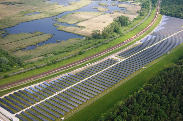 Luftaufnahme eines Solarparks mit Panelen, einem Zug auf einer Schiene, umgeben von Bäumen, Gras, Wasser und Vegetation.