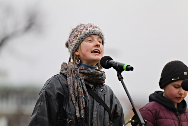 Eine Frau singt in ein Mikrofon auf der Bühne, gekleidet in eine schwarze Jacke, einen Schal und eine Mütze, neben einer Person in einer maronenfarbenen Jacke und schwarzer Mütze.