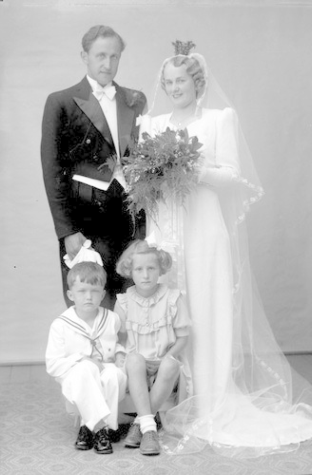 Schwarzes und weißes Foto einer Braut in einem weißen Kleid mit einem Bukett, strahlender Bräutigam neben ihr und zwei Kindern, die vor ihnen auf dem Boden sitzen.