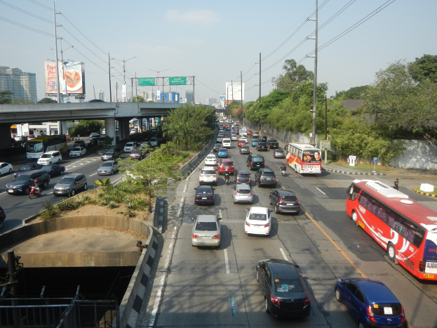 Eine verstopfte Stadtstraße mit Autos, Bussen, Strommasten, Bäumen, Gebäuden, Werbetafeln und einer Brücke unter einem bewölkten Himmel.