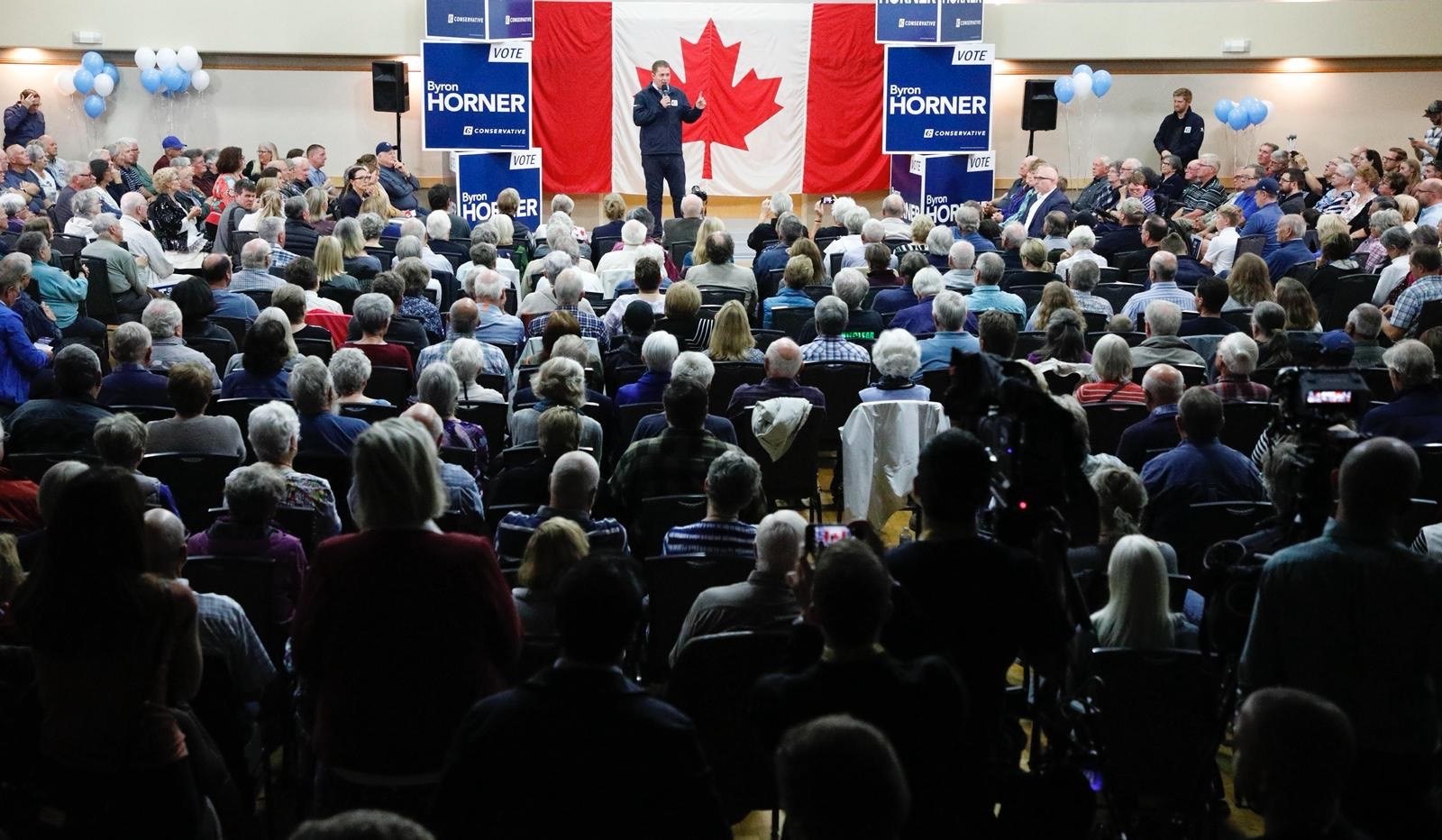 Eine große Menschenmenge sitzt auf Stühlen vor einer kanadischen Flagge, ein Mann hält ein Mikrofon auf einem Podium, umgeben von Bannern, Ballons und Lautsprechern auf dem Byron Horner Conference.