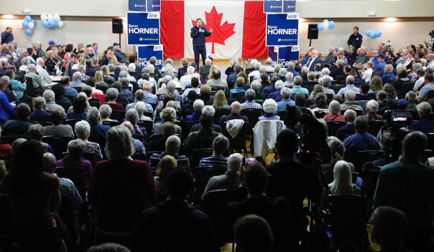 Eine große Menschenmenge sitzt auf Stühlen vor einer kanadischen Flagge, ein Mann hält ein Mikrofon auf einem Podium, umgeben von Bannern, Ballons und Lautsprechern auf dem Byron Horner Conference.