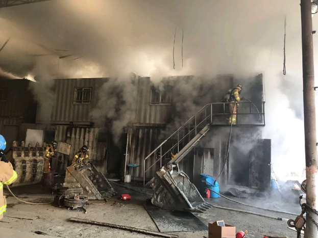 Feuerwehrleute in Helmen und Schutzausrüstung sprühen Wasser, um ein Lagerfeuer zu löschen, mit Rauch, der aus dem Gebäude aufsteigt, Stufen, die zur Tür führen, ein Kettenstab, verstreute Trümmer und Fenster im Hintergrund.
