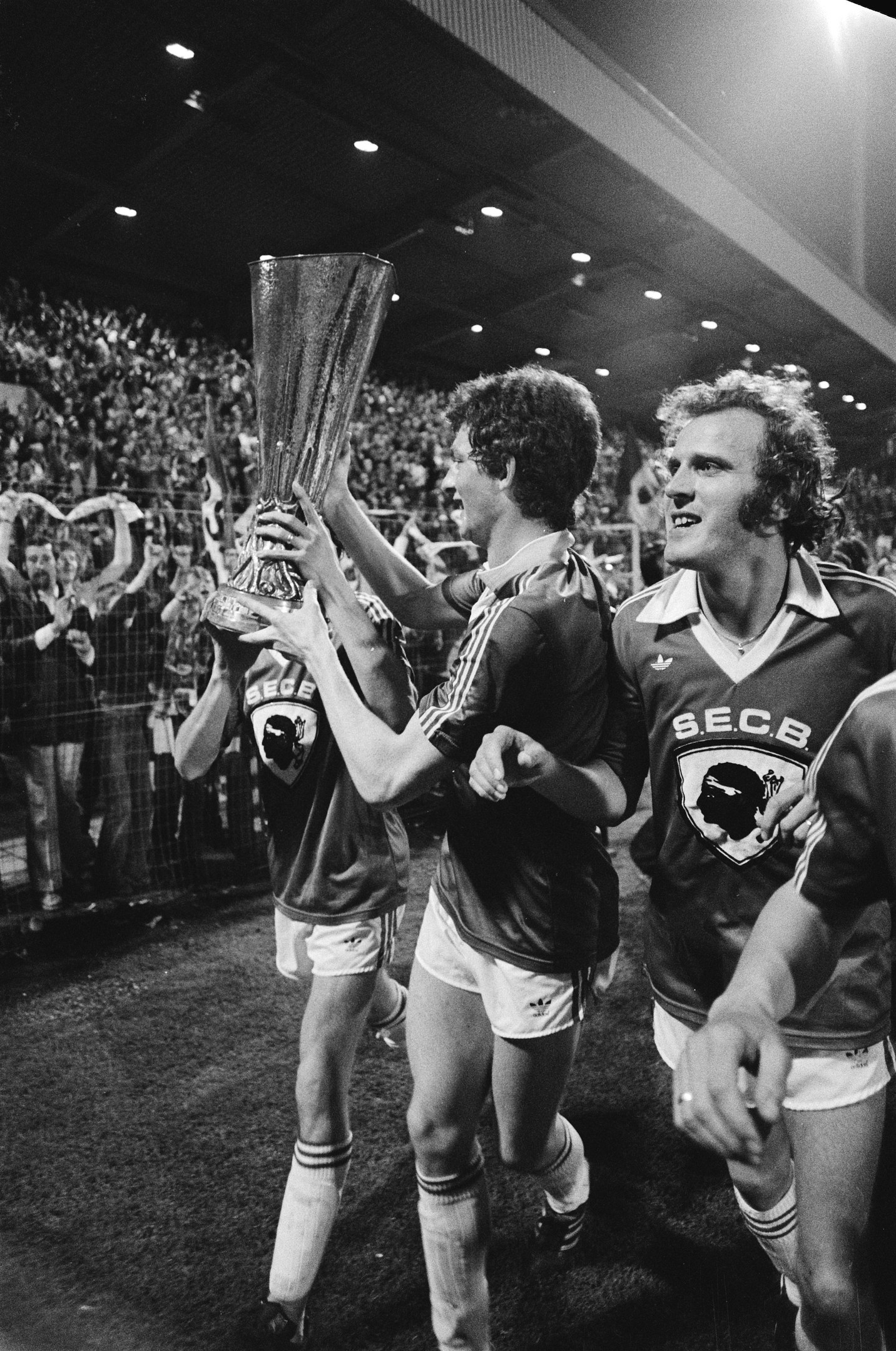 Schwarzes Foto von Fußballspielern, die einen Pokal auf einem Feld halten und ihren Sieg in der FA Cup Final feiern, während Zuschauer hinter einem Zaun zusehen.