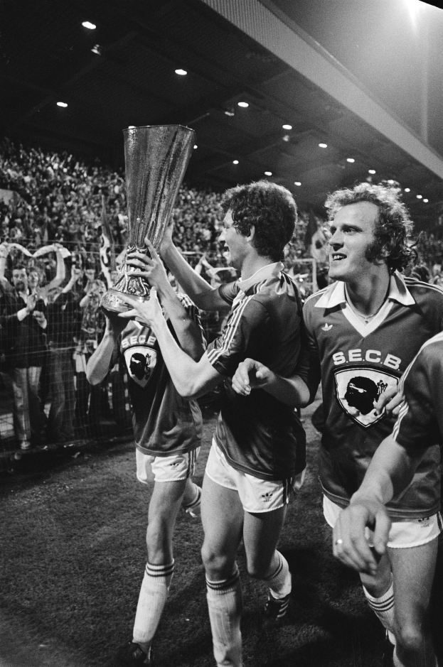 Schwarzes Foto von Fußballspielern, die einen Pokal auf einem Feld halten und ihren Sieg in der FA Cup Final feiern, während Zuschauer hinter einem Zaun zusehen.