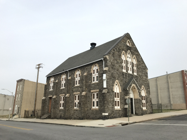 Alte steinerne Kirche an einer Straßenecke mit umgebenen Gebäuden, Straßeninfrastruktur und Fahrzeugen unter einem klaren blauen Himmel.