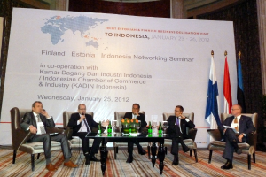 Eine Konferenzbühne mit Menschen, Stühlen, einem Mikrofon, Kabeln, einem Tisch, Flaschen, Blumen, Gläsern und anderen Gegenständen, mit Flaggen und einem Banner in der Mitte und einem Gebäude mit Vorhängen im Hintergrund.