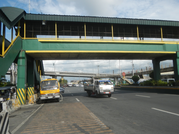 Mautstelle mit Fahrzeugen darunter, Brücke mit Geländern und Säulen, Laternenmasten, Schilder, Bäume und bewölkter Himmel im Hintergrund, mit einem Lastwagen, der neben der Brücke fährt.