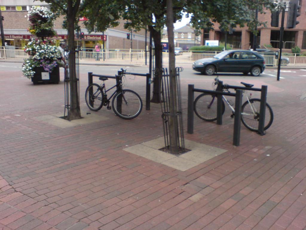 Eine Szene mit Fahrrädern, Bäumen, einem Zaun, einem Auto und Gebäuden.