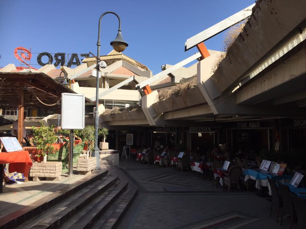Eine Straße mit Tischen und Stühlen in der Mitte, umgeben von Gebäuden, Straßenmöbeln, Pflanzen und einem klaren blauen Himmel, mit Büchern und anderen Gegenständen auf den Tischen.