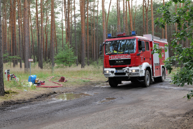Rotes Feuerwehrauto fährt auf einer Schotterstraße mit Bäumen auf beiden Seiten, Gras und ein Rohr auf der linken Seite.