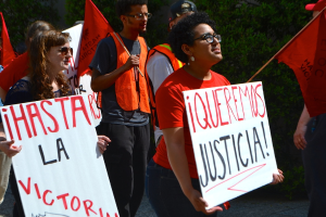 Mehrere Personen in orangefarbigen Kleidern halten weiße Schilder und orangefarbene Fahnen, mit einer Frau in einem schwarzen Kleid im Vordergrund und grünen Bäumen im Hintergrund.