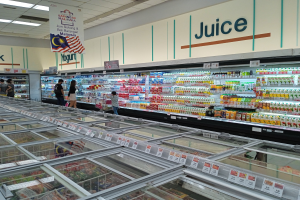 Ein gut sortierter Supermarktwarengang mit Regalen voller Getränkeflaschen, Obst und anderen Lebensmitteln, einige Einkäufer und Deckenbeleuchtung.