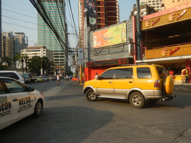 Ein gelbes Taxi fährt auf einer Straße mit hohen Gebäuden, Passanten auf dem Gehweg, Strommasten mit Drähten, Bäumen und einem sichtbaren Himmel.
