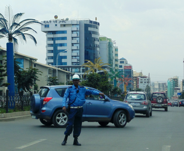Polizist in blauer Uniform und weißem Helm vor einem Auto auf einer Stadtstraße mit Gebäuden, Bäumen und Fahrzeugen im Hintergrund.