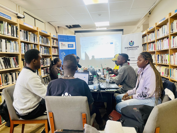 Gruppe von Menschen, die um einen Tisch in einer Bibliothek mit Bücherregalen, Laptops, Flaschen und Taschen sitzen und über eine Wikimedians- und Kamerun-Initiative mit einem Bildschirm und Bannern im Hintergrund diskutieren.