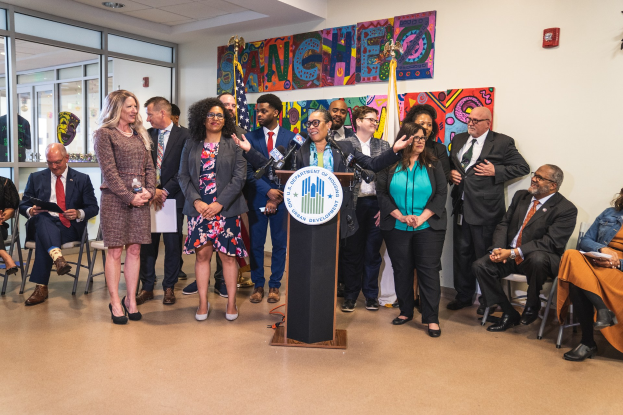 Eine Gruppe von Menschen steht vor einem Rednerpult mit einem Mikrofon und einer Tafel mit der Aufschrift "Sancho-Stadtrat tritt dem Stadtrat bei", im Hintergrund sind weitere Personen auf Stühlen und auf dem Boden sitzend und stehend zu sehen, sowie Flaggen, eine Tafel, einen Feuerlöscher und Deckenleuchten an der Wand.