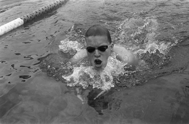 Ein Mann schwimmt in einem Pool während eines Rennens, trägt eine Schwimmbrille und ist von Bahnmarkierungen umgeben, dargestellt in Schwarz-Weiß.