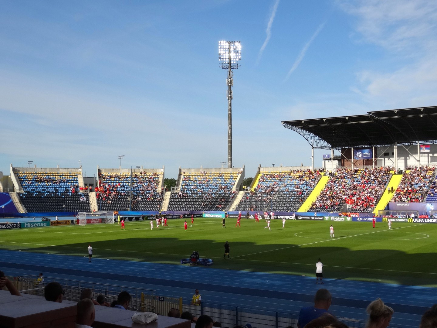 Ein Fußballspiel in einem großen Stadion mit Spielern auf dem Feld und Zuschauern in den Rängen unter Flutlicht, mit dem Himmel im Hintergrund.