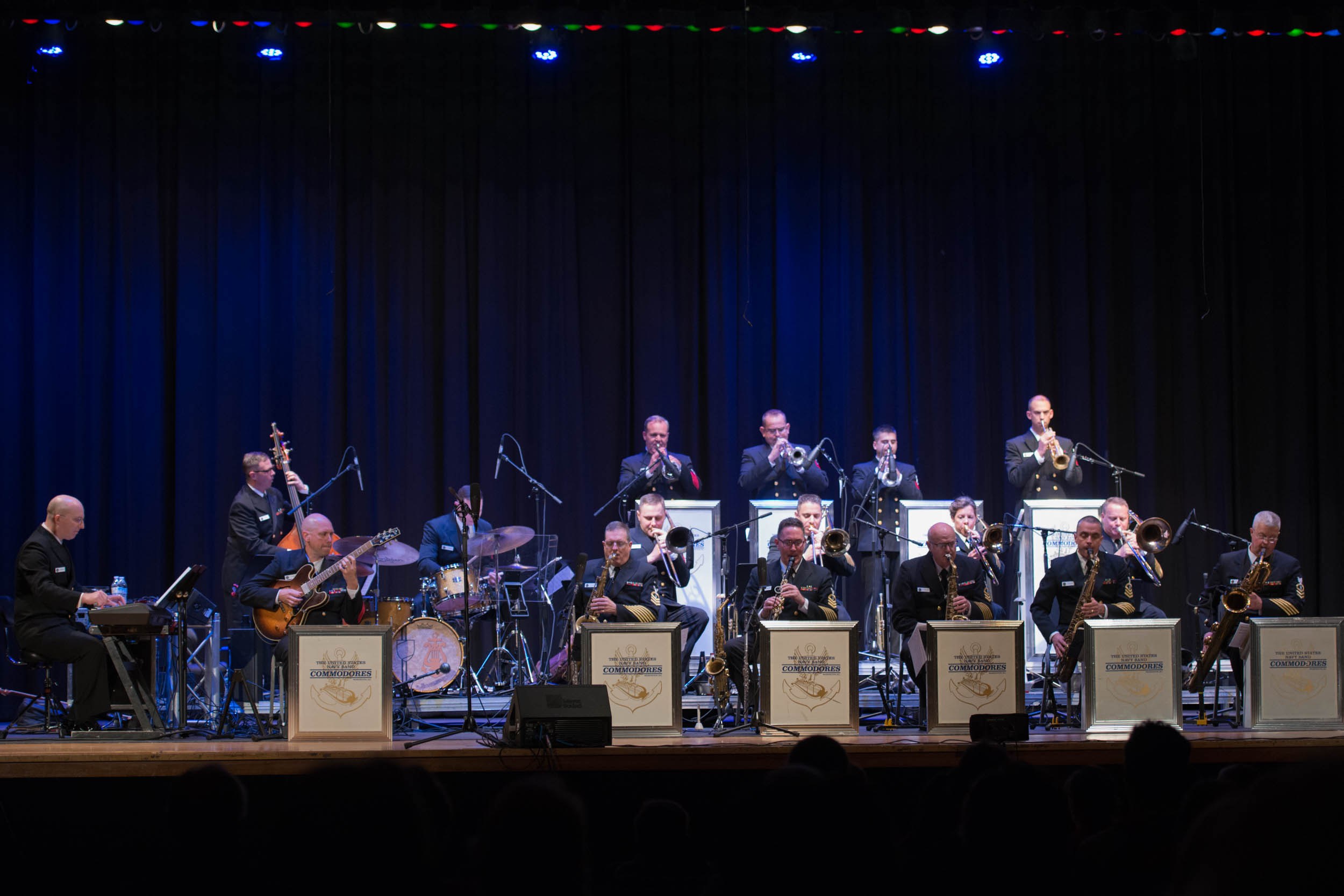 Eine Gruppe von Musikern tritt auf einer hell erleuchteten Bühne auf, die mit Mikrofonen, Ständern und Texttafeln versehen ist, während das Publikum von unten zusieht.