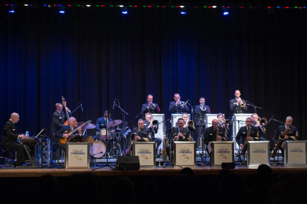 Eine Gruppe von Musikern tritt auf einer hell erleuchteten Bühne auf, die mit Mikrofonen, Ständern und Texttafeln versehen ist, während das Publikum von unten zusieht.