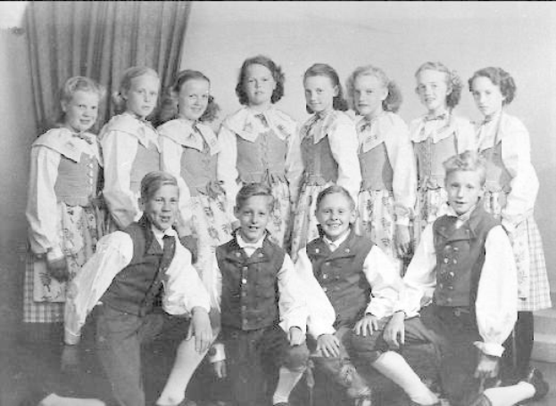 Schwarzes und weißes Foto einer Gruppe von Kindern, die vor einer Vorhangwand posieren, mit dem Text "1930er-Kinder-Schule" unten.