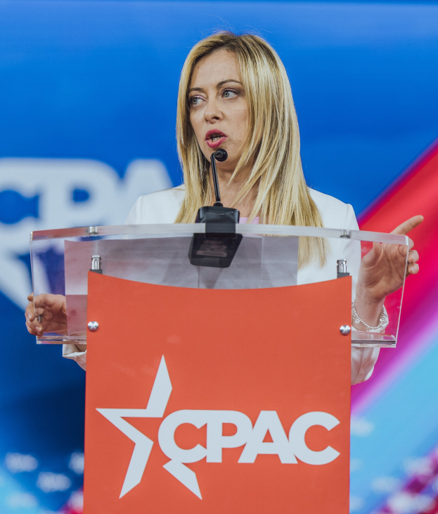 Frau in weißem Kleid, die an einem Rednerpult mit Mikrofon spricht, unscharfer Hintergrund, wahrscheinlich eine Politologin.