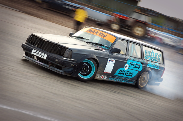 Ein Auto, das auf einer Rennstrecke fährt und eine Rauchspur hinterlässt, umgeben von anderen Fahrzeugen und Zuschauern.