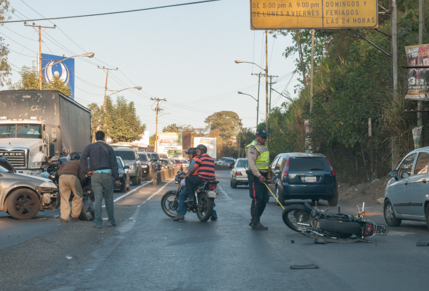 Gruppe von Menschen versammelt um ein verunglütztes Motorrad am Straßenrand mit mehreren Fahrzeugen, darunter ein Lastwagen, im Hintergrund und Bäumen, Masten, Lichter, Bretter und Himmel.
