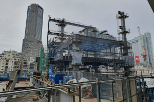 Ein großes Gebäude im Bau in einer Stadt, umgeben von einem Metallzaun, mit benachbarten Gebäuden, Polen, einer Fahne, Fahrzeugen, Drähten und einem bewölktem Himmel.