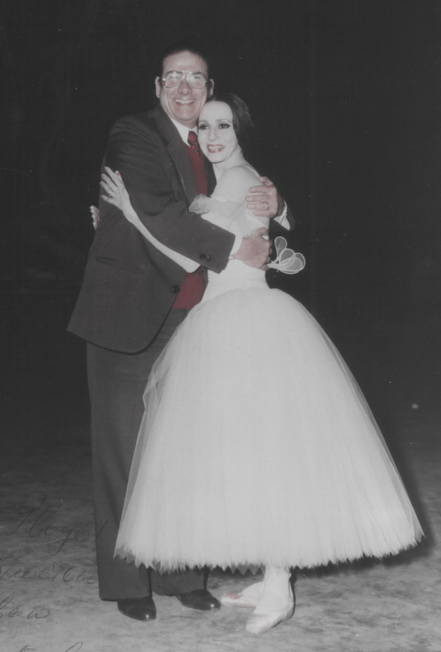 Schwarzes und weißes Foto eines Mannes in Blazer, Krawatte, Hemd und Hose, der eine Frau in Schuhen umarmt, mit einem dunklen Hintergrund, der eine romantische Atmosphäre schafft.