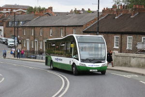 Ein grüner und weißer Bus fährt auf einer Straße mit hohen Gebäuden, Fußgängern auf dem Gehweg, Radfahrern, Geländern, Treppen, Straßenschildern, Bäumen und einem klaren blauen Himmel.