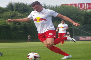 Ein professioneller Fußballspieler schießt den Ball auf einem Rasenfeld mit Bäumen und einem klaren blauen Himmel im Hintergrund, wobei ein "RB Leipzig"-Schild sichtbar ist.